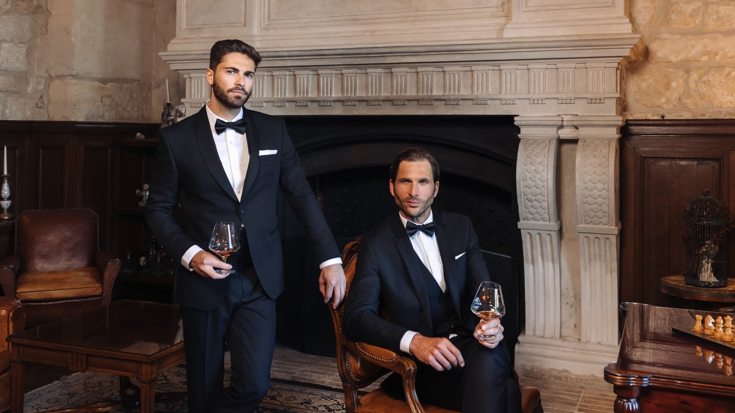 Two guests in dinner jackets beside the carved stone fireplace of the Cognathèque