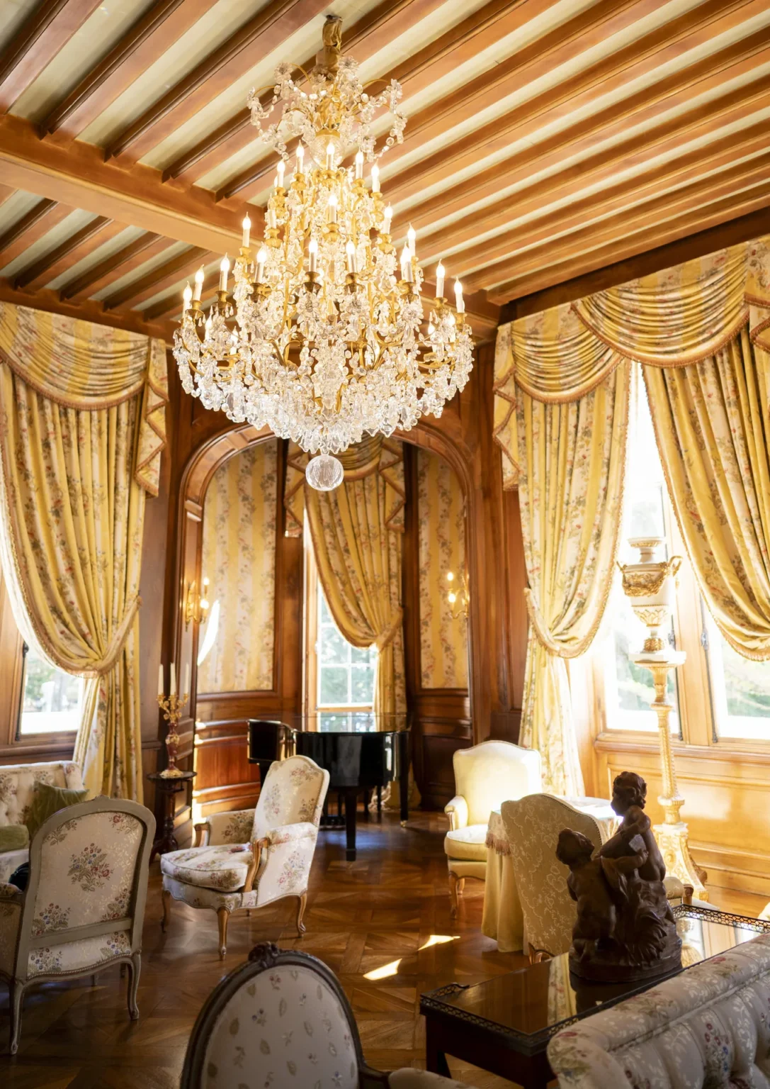 Grand Salon with a crystal chandelier, tall draped windows, armchairs and a grand piano