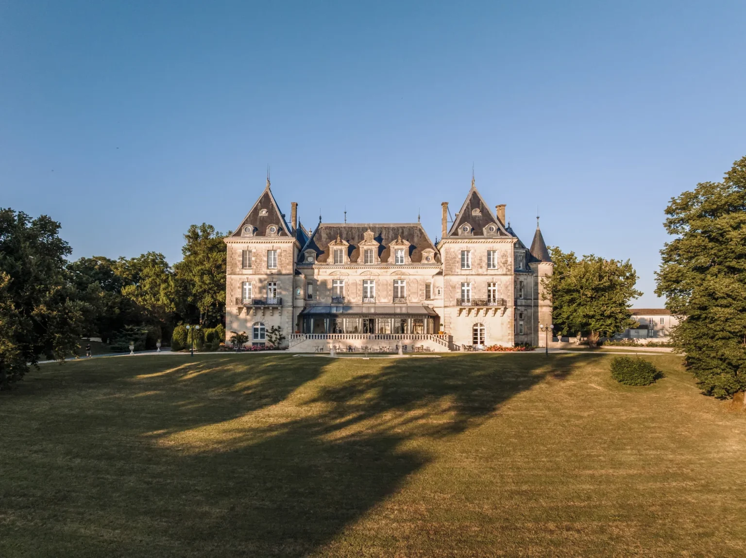 Le château de Mirambeau, vu depuis le pelouse en lumière de soirée, avec sa façade complète et ses tours recouvertes d’arbres