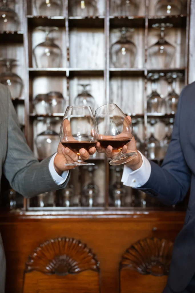 Two guests raising glasses of cognac in front of a wooden cabinet of glassware