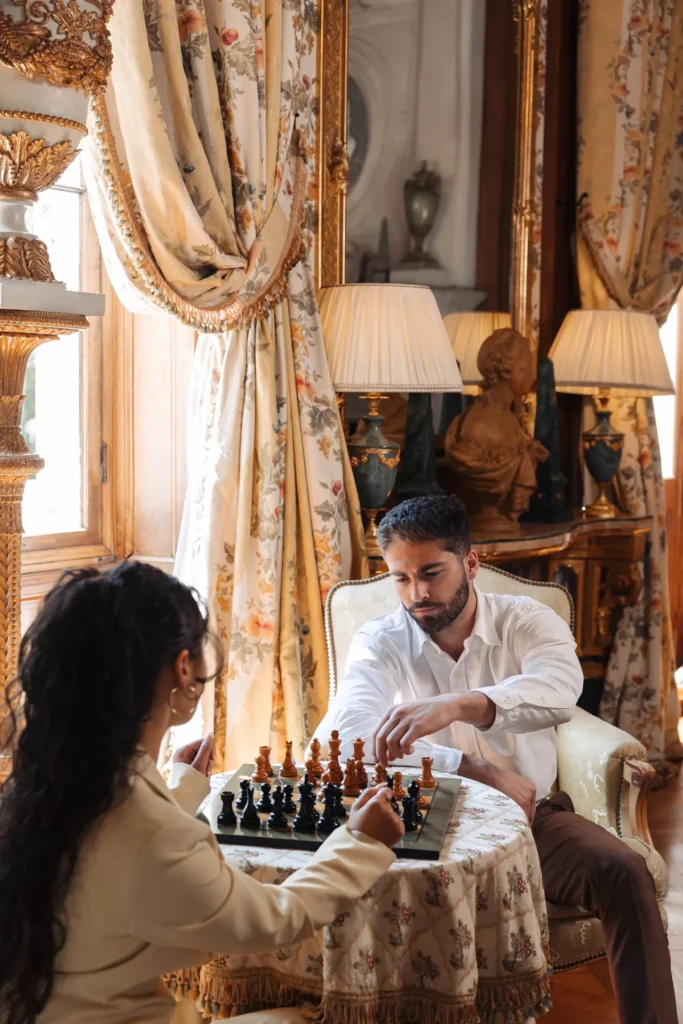 Two guests playing chess at a small table in a salon with floral curtains