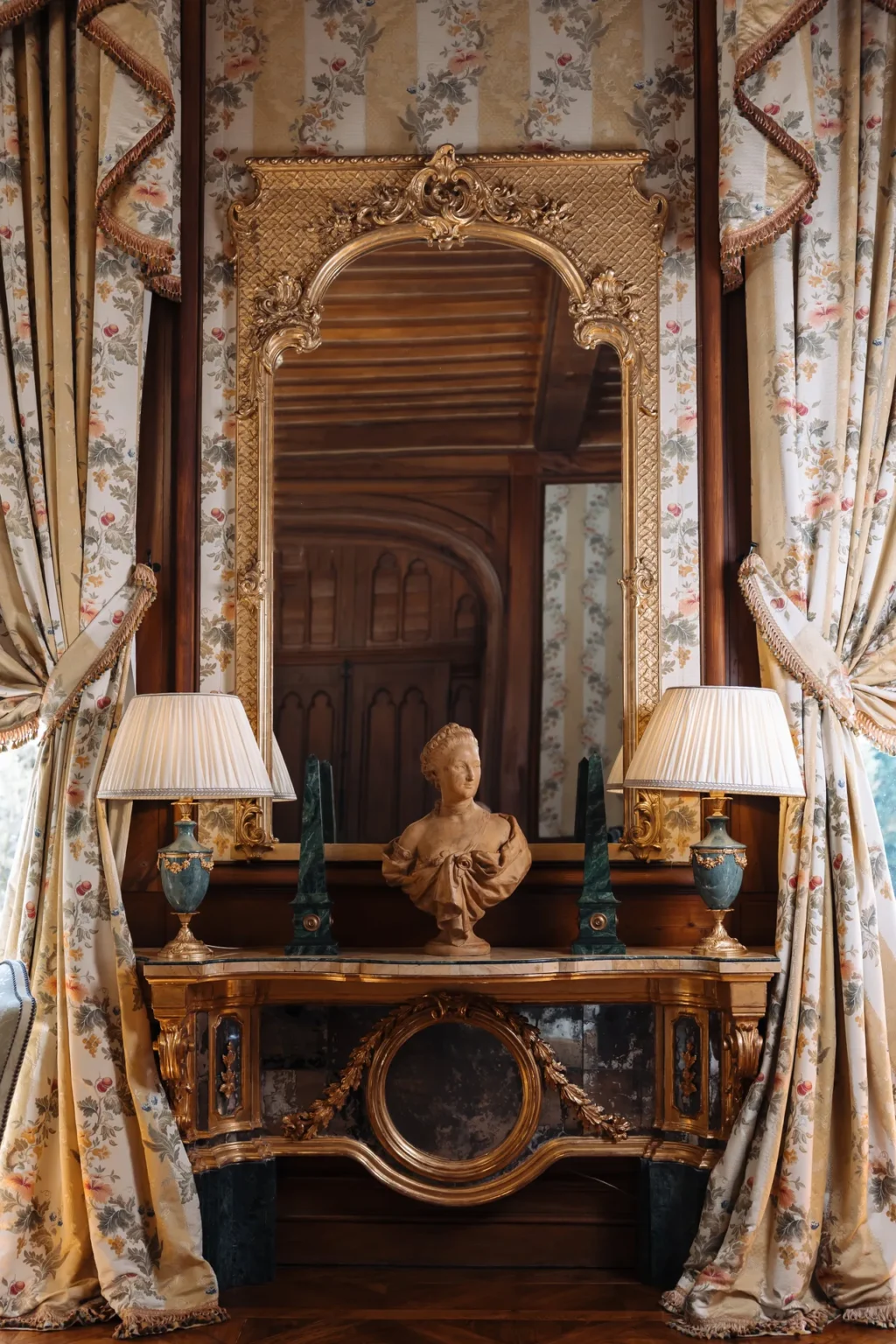 Carved wooden doorway and gilded console with lamps and a bust, framed by floral curtains in the château