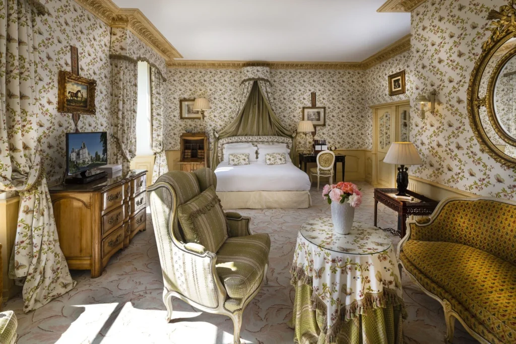 A suite with a canopied bed, armchairs and a skirted round table beneath floral wallpaper