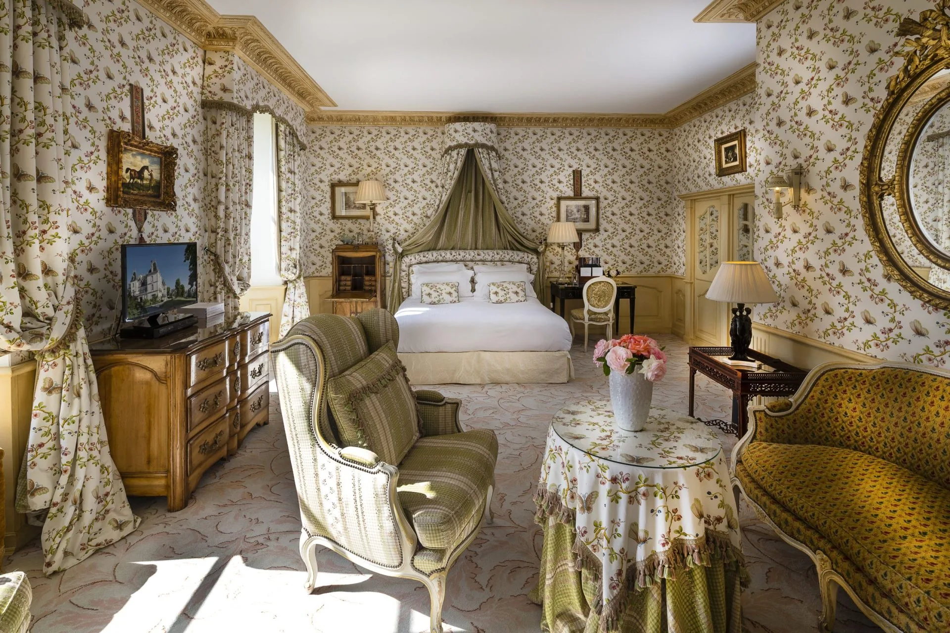A suite with a canopied bed, armchairs and a skirted round table beneath floral wallpaper