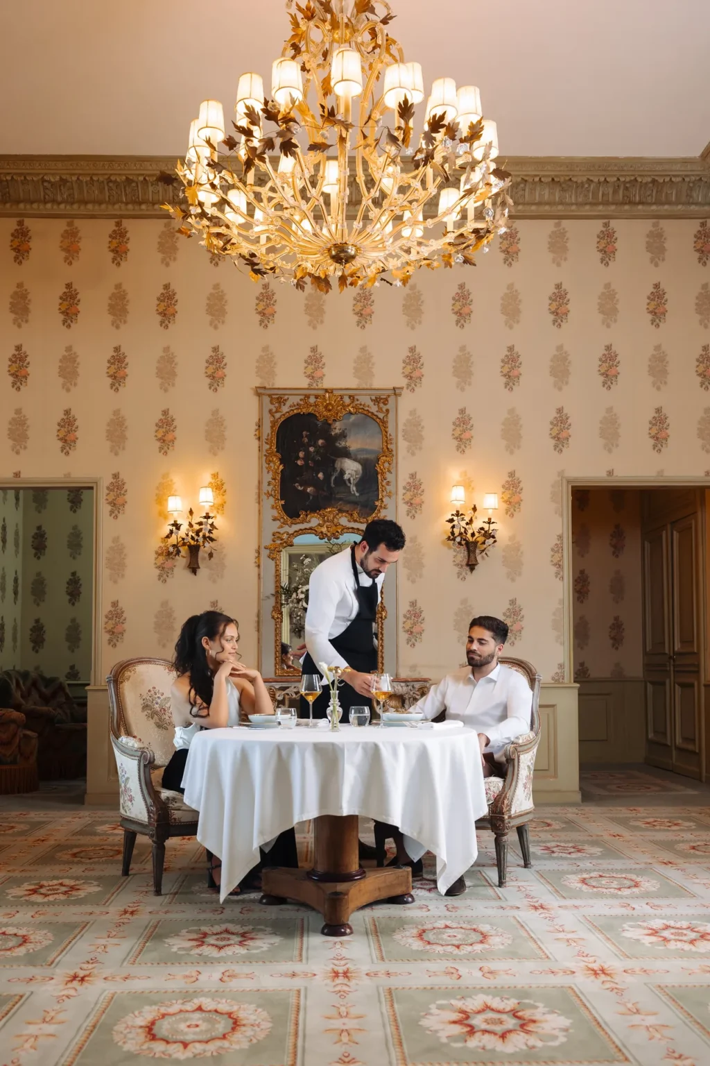 Two guests dining in a salon beneath a chandelier while a waiter serves