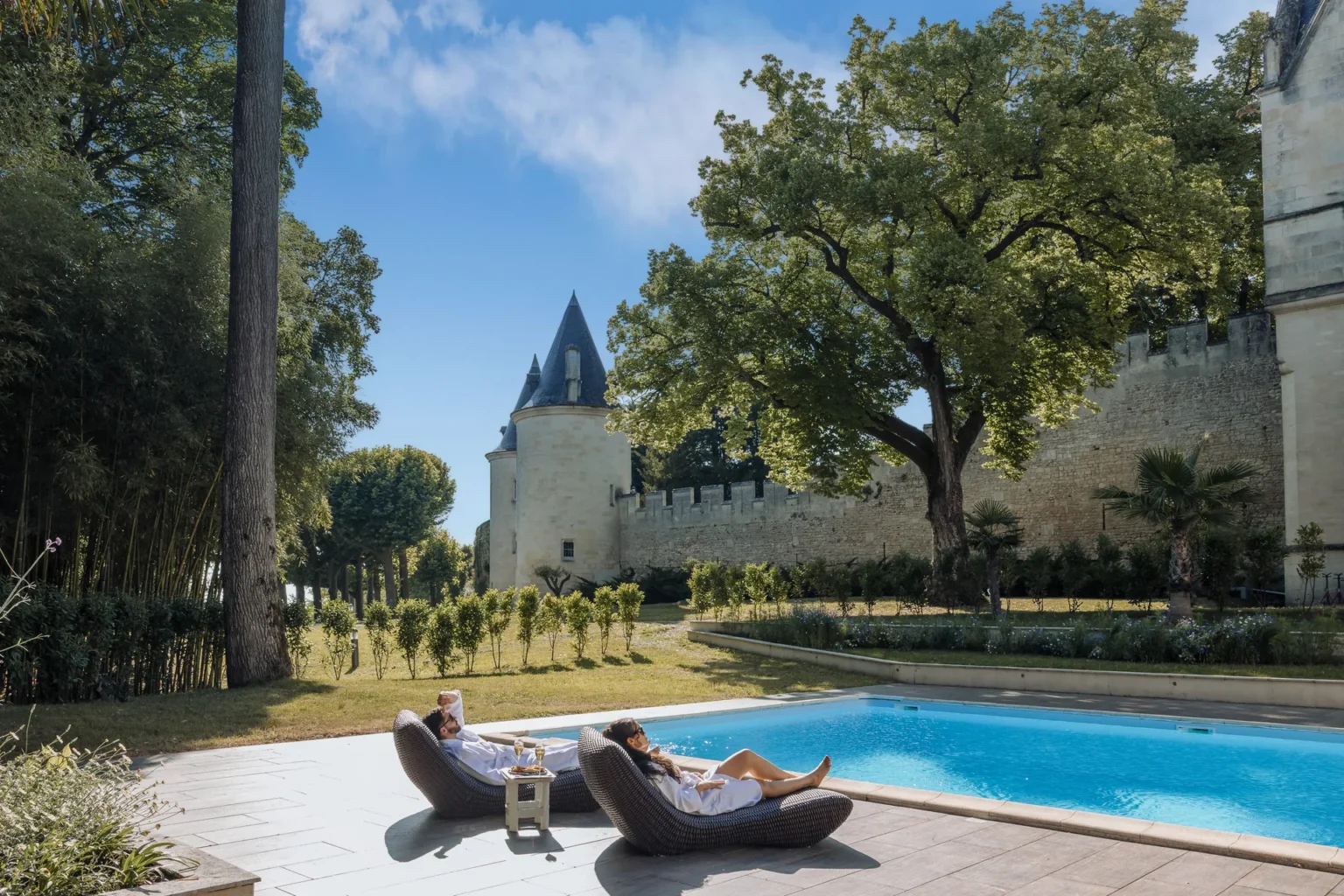 Guests on loungers by the pool with a château tower and trees in the background
