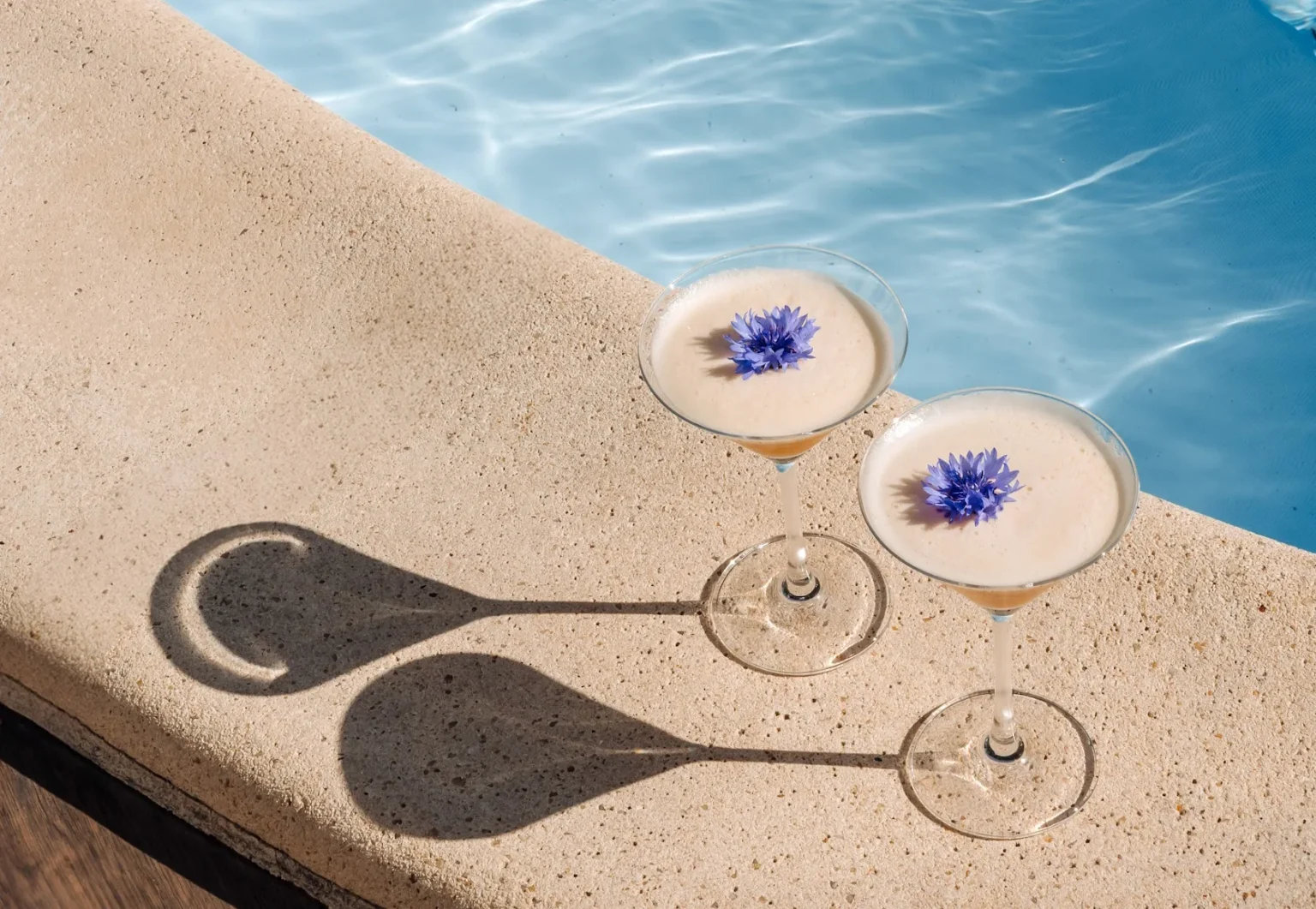 Two cocktails garnished with cornflowers on the stone edge of the pool