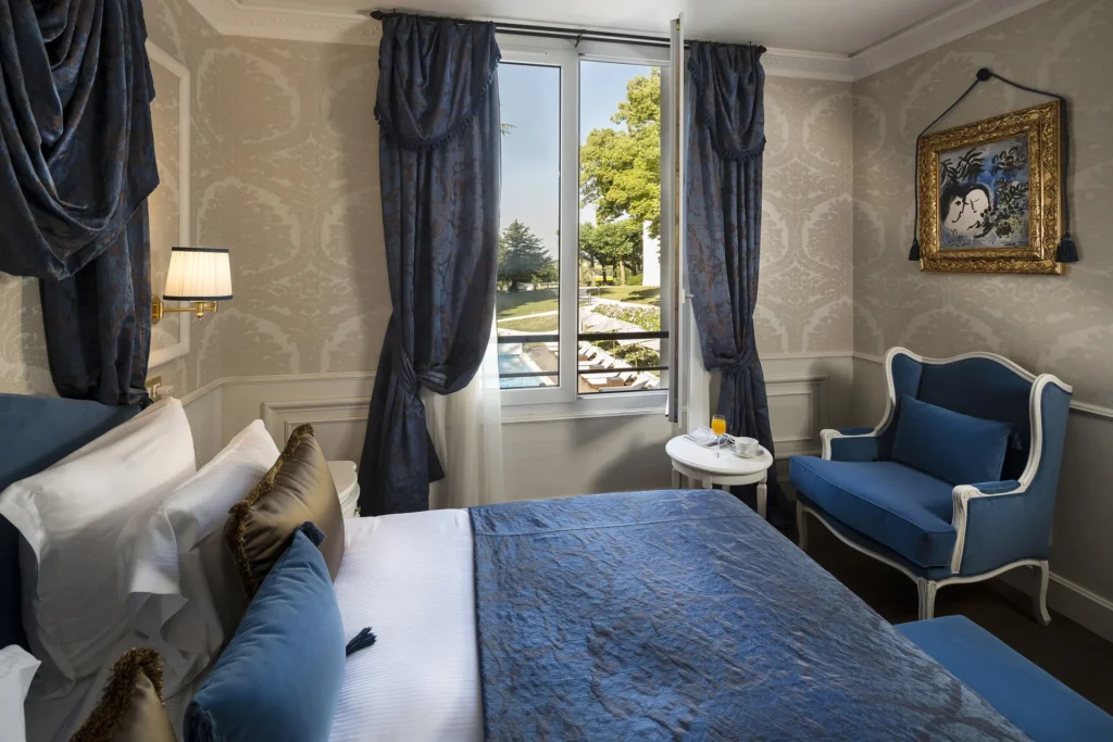 A Superior Room in the Orangerie with a blue armchair and a window looking out towards the pool