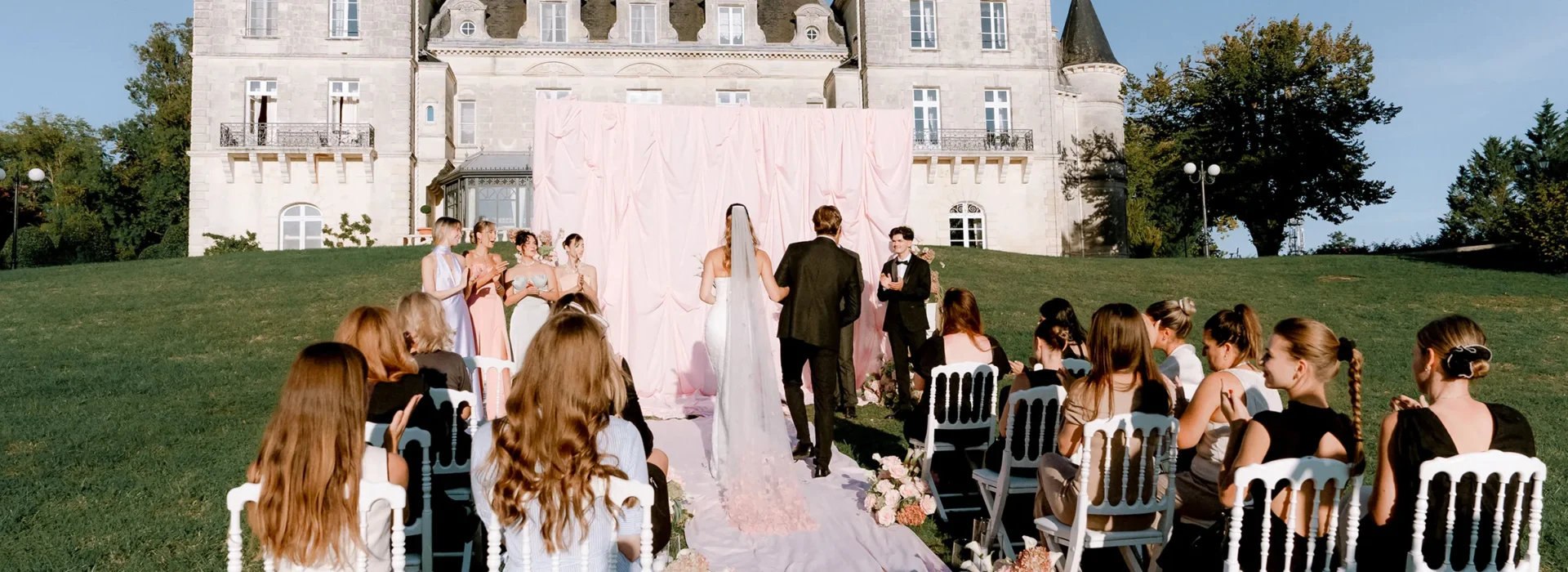 Guests gathered on the lawn before the château façade around the couple beneath a flower arch